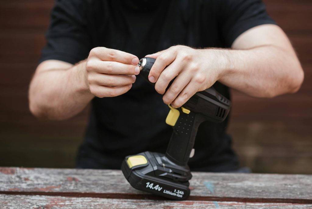 Crop anonymous skilled master in casual outfit preparing electric drill for job on wooden table in soft daylight